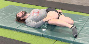 Dr. John trying to keep both shoulders flat on the ground while pulling his left leg across his body with his right arm and his right leg bent in a V position to stretch his hip flexor and quad